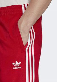 Červené sportovní šortky z texturované látky, s bílými třemi pruhy a malým bílým logem Adidas na levé straně.