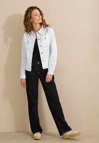 Vitt denimjacka med två bröstfickor, i kombination med mörka jeans med gula sömmar och ljusa sneakers.