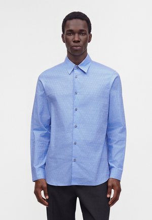 Homme portant une chemise bleu clair à manches longues avec des motifs subtils, debout devant un fond blanc uni.