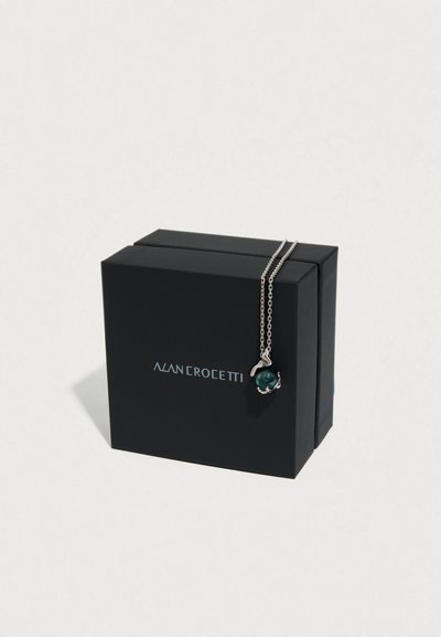 Ασημένιο κολιέ με μενταγιόν από πράσινη πέρλα, τοποθετημένο σε μαύρο κουτί με λογότυπο. Απλός σχεδιασμός αλυσίδας που προσθέτει κομψότητα.