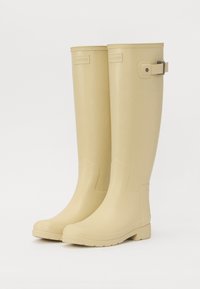 Botas de lluvia de goma beige con punta redondeada, altura hasta la mitad de la pantorrilla, borde superior texturizado y detalle de hebilla ajustable en el lateral.