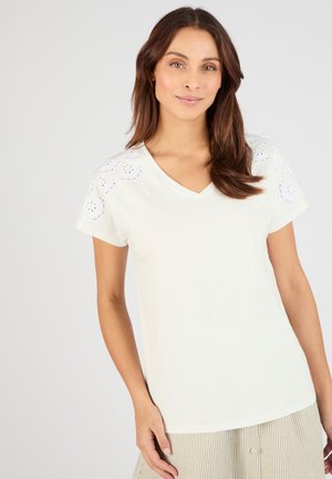 Vrouw met bruin schouderlang haar, draagt een wit T-shirt met V-hals en een uitgewerkt bloemenpatroon op de mouwen en een gestreepte beige rok.