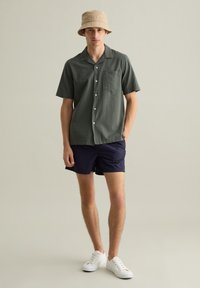 Camicetta verde con maniche corte e texture, dotata di tasca, abbinata a pantaloni corti blu navy e scarpe da ginnastica bianche. Un cappello a secchiello color sabbia completa il look.