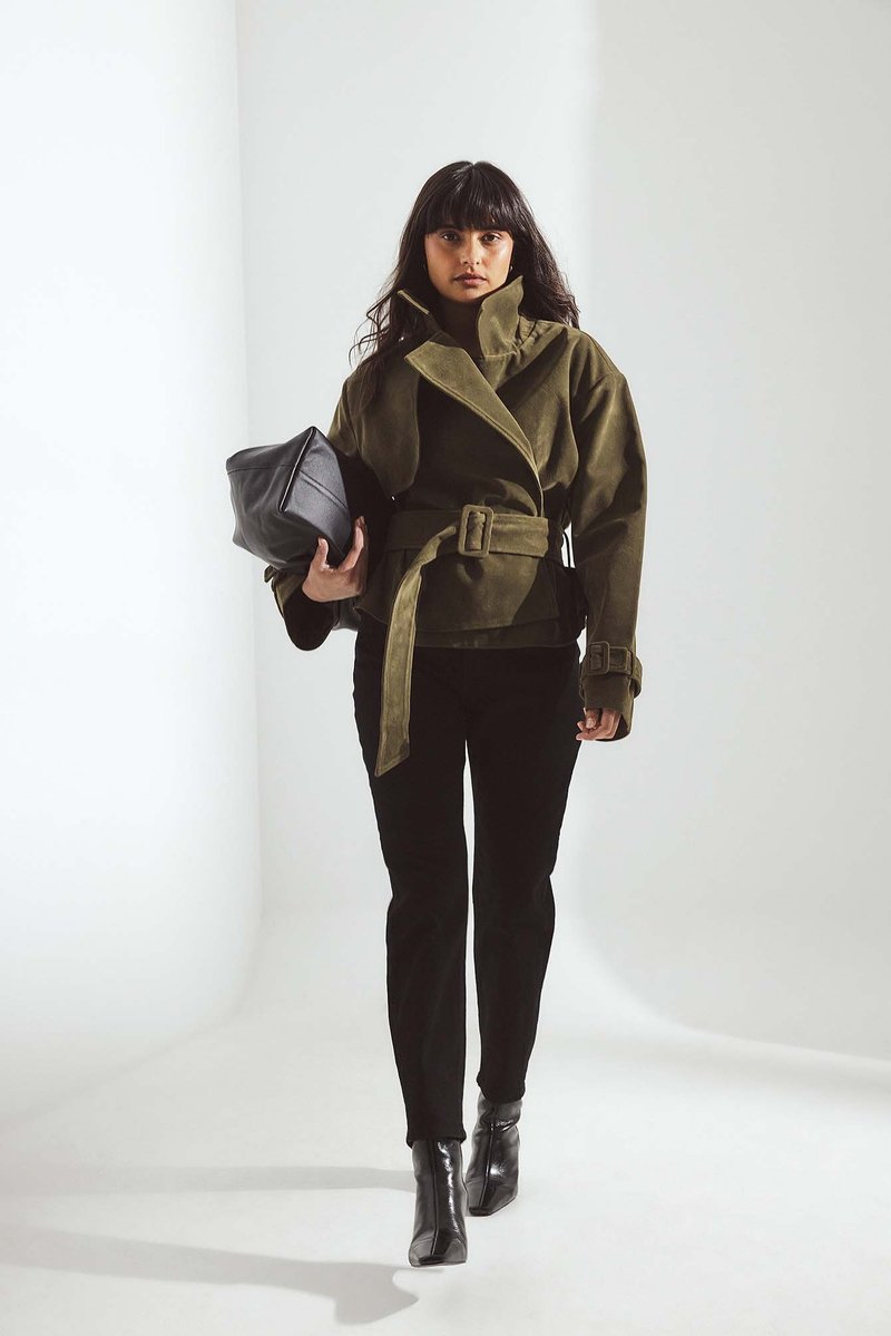 Chaqueta verde oliva con cinturón y cuello alto, hecha de tela texturizada; combinada con pantalones negros ajustados y botas de tacón. Bolso negro en la mano.