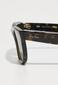 Ochelari de soare Ray-Ban din material tartan, cu un cadru gros, având brandul argintiu, un design unghiular și lentile gri. Fabricat în Italia.
