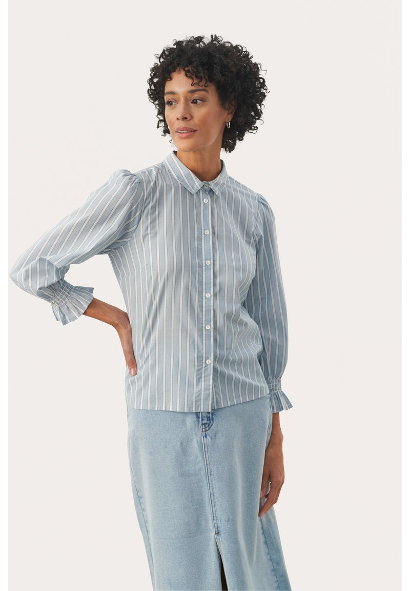 Lichtblauw gestreept overhemd met opgeblazen mouwen, afgeronde kraag en gerimpelde manchetten, gecombineerd met een denim rok met een voorkuipe.