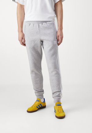 Mężczyzna ubrany w jasnoszare spodnie dresowe Adidas z białymi paskami i żółte buty sportowe Adidas z niebieskimi paskami na białym tle.