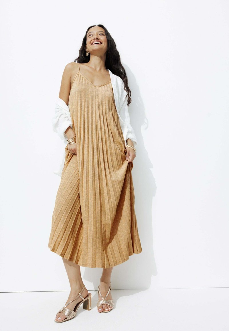 Robe midi plissée dorée avec un décolleté en V et des fines bretelles, accompagnée d'un cardigan blanc et de sandales à talons bloc beiges.