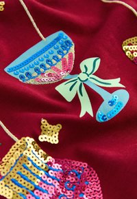 Rødt stof med farverige pailletdekorationer i form af kopper, sløjfer og mønstre. Inkluderer detaljer i guld, pink, blå og grøn.