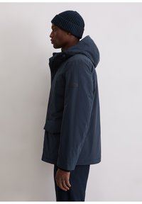 Marineblauwe waterdichte parka met capuchon, voorzien van een voorflappzak, verstelbare manchetten en een gladde afwerking. Draagt een donker gebreide muts.