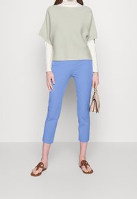 Ljusgrön texturerad stickad topp, vit långärmad tröja under; blå figurnära ankelbyxor; bruna platta sandaler; håller en liten tan handväska.