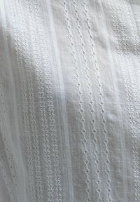 Valkoinen kangas, jossa on monimutkaisia brodeerattuja kuvioita, geometaalisia muotoja, suoria viivoja ja teksturoituja yksityiskohtia. Kevyt ja ilmava ilme.