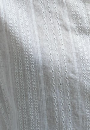 Bijeli materijal s složenim izvezenim uzorcima koji sadrže geometrijske oblike, ravne linije i teksturirane naglaske. Lagani i prozračni izgled.