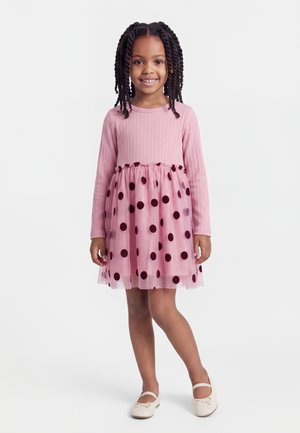 Fille aux cheveux tressés portant une robe rose à manches longues avec une jupe à pois et des chaussures blanches, debout et souriante sur un fond blanc.