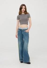Top grigio corto a costine abbinato a jeans in denim blu a gamba larga con frange verdi lungo i lati. Calzature nere completano il look.