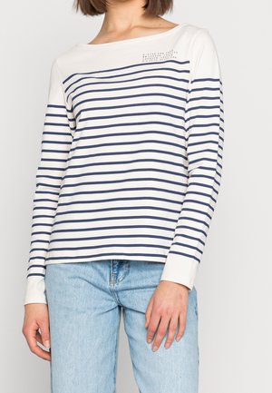 Mujer con camisa blanca de manga larga con rayas horizontales azul marino y jeans azul claro, de pie frente a un fondo blanco liso.