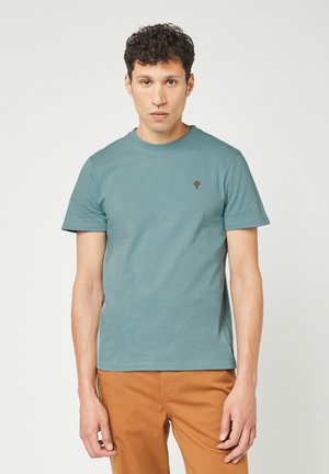 Korte mouw teal t-shirt van katoen met een ronde halslijn en een klein bruin boompje logo op de linkerborst, gecombineerd met beige broek.