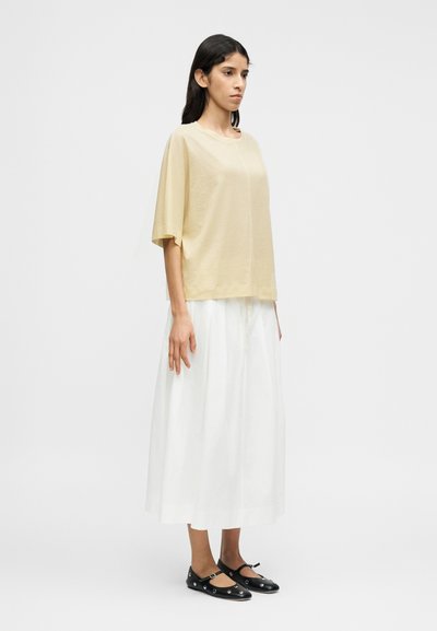 Κοντομάνικο, ανοιχτό κίτρινο τοπ από απαλό ύφασμα, σε συνδυασμό με μακριά λευκή φούστα και μαύρα επίπεδα παπούτσια με διακοσμητικά αγκράφες.