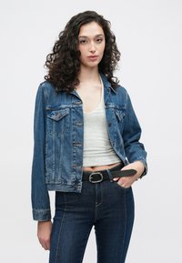 Modra denim jakna s krajšim krojem, z gumbi spredaj, dvema žepoma na prsih in rahlim bledenjem. Oblečena čez sivo majico in temne kavbojke.