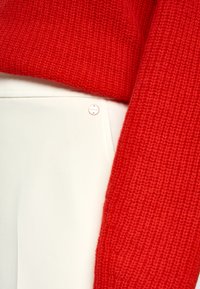 Roter Strickpullover liegt über cremefarbenen Hosen. Kleine goldfarbene Metallakzente sind an der Hosentaschennähte sichtbar.