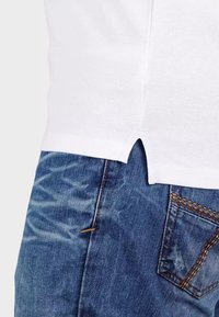 T-shirt bianca in cotone con spacchi laterali, abbinata a jeans in denim blu con texture sbiadita e cuciture arancioni sui taschini.