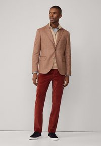 Brauner Fischgrät-Blazer über einem beigen Pullover, kombiniert mit roten Cordhosen und marineblauen Sneakers. Ordentliche, strukturierte Details mit einer taillierten Passform.
