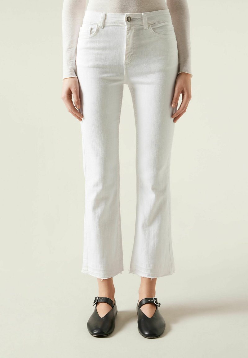 Jeans de mezclilla blanca acampanados con dobladillo sin terminar, diseño de cinco bolsillos y cierre de cremallera. Combinados con zuecos de cuero negros.