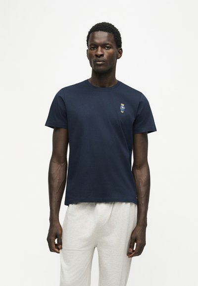 Homme portant un t-shirt bleu marine avec un petit ours brodé sur la poitrine et un pantalon gris clair, debout devant un fond blanc uni.