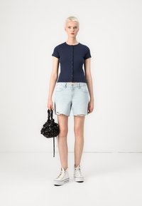 Haut à manches courtes côtelé marine avec fermeture à boutons, short en denim bleu clair effiloché, baskets montantes blanches et mini sac noir à motifs.
