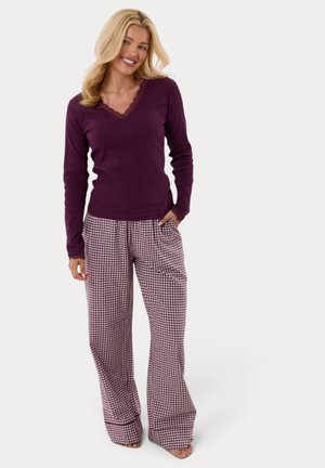 Mujer de pie sonriendo, vistiendo una camiseta morada de manga larga y pantalones anchos de cuadros con una mano en el bolsillo.