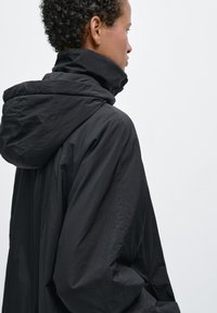 Giacca nera con cappuccio dal taglio comodo, caratterizzata da un collo alto e un tessuto morbido e leggero, che presenta una texture liscia e un design minimalista.