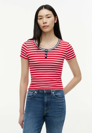 Junge Frau mit langen schwarzen Haaren, die ein rot-weiß gestreiftes kurzärmliges Oberteil und blaue Jeans trägt, vor einem weißen Hintergrund stehend.