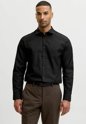 Homme portant une chemise noire à manches longues boutonnée et un pantalon marron, debout devant un fond clair uni.
