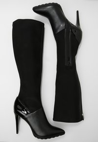 Botas negras por encima de la rodilla que combinan ante y charol, con punta estrecha, tacón alto de aguja y detalle de cremallera lateral.