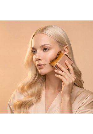 Blonde woman with wavy hair applies makeup with a wide, curved brush on her cheek against a beige background.