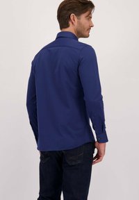 Man in een donkerblauw overhemd met lange mouwen en donkere jeans, staande met de rug naar de camera tegen een effen lichte achtergrond.