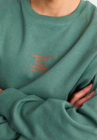 Zielony sweatshirt z okrągłym dekoltem, z czerwonym nadrukiem zawierającym napisy "TIMBERLAND" i "EST 1973". Miękka faktura, długie rękawy, luźny krój.