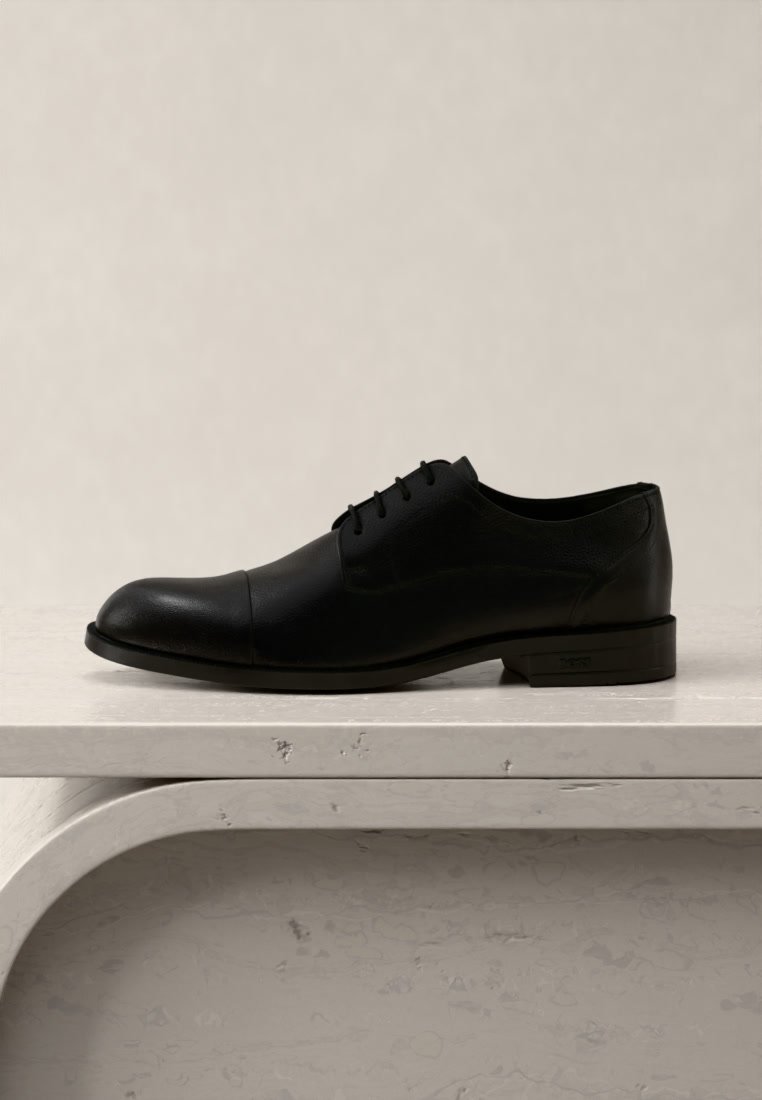 Chaussure habillée formelle en cuir noir avec des lacets, posée sur une surface en pierre texturée de couleur claire, sur un fond beige uni.