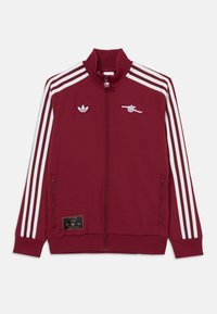 Bordová zippová bunda s vysokým golierom, biele trojpruhové akcenty, logo Adidas a emblém Arsenalu. Obsahuje bočné vrecká a ribované manžety.