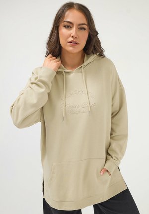 Frau trägt einen beigen, übergroßen Hoodie mit der Stickerei "Palm Springs Tennis Club California", posiert mit einer Hand nahe der Kapuze und der Tasche.