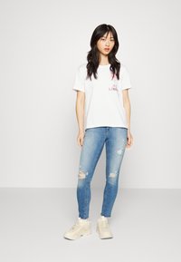 White cotton t-shirt with short sleeves and a graphic print at the chest, paired with light blue distressed skinny jeans and beige sneakers.