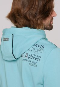 Light blue hooded sweatshirt made of soft fabric. Features graphic text designs in dark blue and silver accents on the upper back.