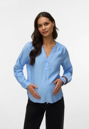 Femme enceinte portant un chemisier à rayures bleues et un pantalon noir, tenant délicatement son ventre avec les deux mains sur un fond uni.