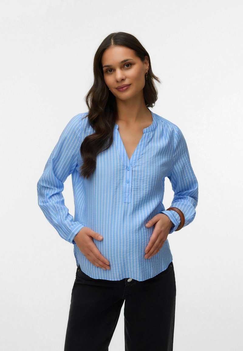 Gravid kvinde iført en blåstribet bluse og sorte bukser, der blidt holder sin mave med begge hænder mod en ensfarvet baggrund.