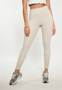 Beige korkeavyötäröiset leggingsit, joissa on sileä pinta ja tiukka muotoilu, yhdistettyinä harmaisiin urheilukenkiin, joissa on valkoiset nauhat ja yksityiskohdat.