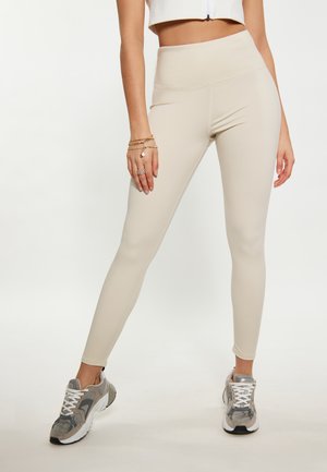Beige korkeavyötäröiset leggingsit, joissa on sileä pinta ja tiukka muotoilu, yhdistettyinä harmaisiin urheilukenkiin, joissa on valkoiset nauhat ja yksityiskohdat.