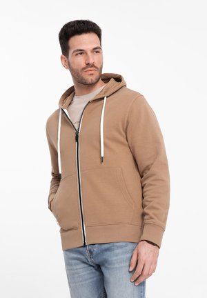 Hooded zip-up sweatshirt, tan color, made from soft fabric. Features white drawstrings, front pockets, and ribbed cuffs. Worn over a light shirt.