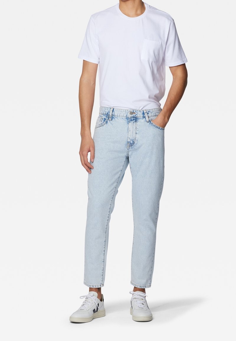 Homme portant un t-shirt blanc uni à manches courtes, un jean bleu clair et des baskets blanches, debout avec une main dans la poche.