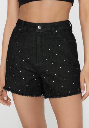 Femme portant un short en denim noir taille haute avec de petites clous argentés et des bords effilochés, associé à un top noir court.