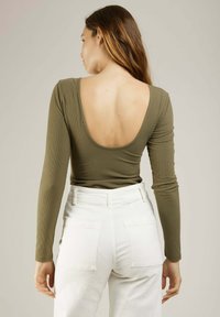 Bodysuit vert olive à manches longues avec un décolleté profond au dos, associé à un pantalon blanc taille haute avec deux poches arrière.